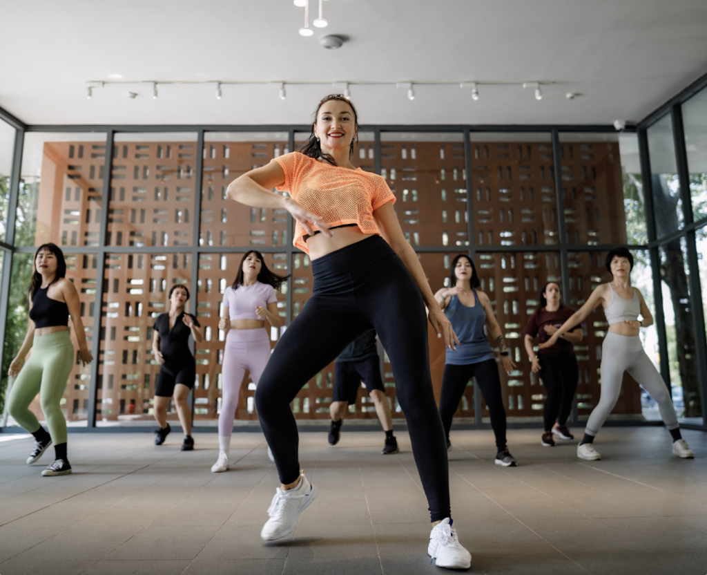 Instructor leads a high-energy dance fitness class in a bright studio, foreground dancer smiling in orange top and black leggings with peers behind her in colorful athletic wear.