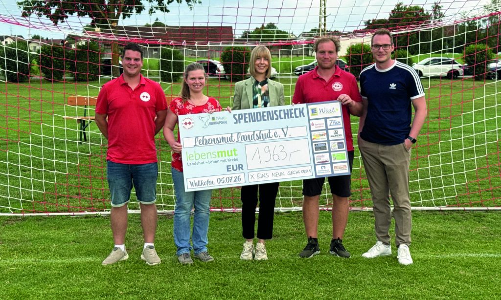 Five people stand in front of a soccer goal holding a large donation check, smiling on a sunny grassy field.