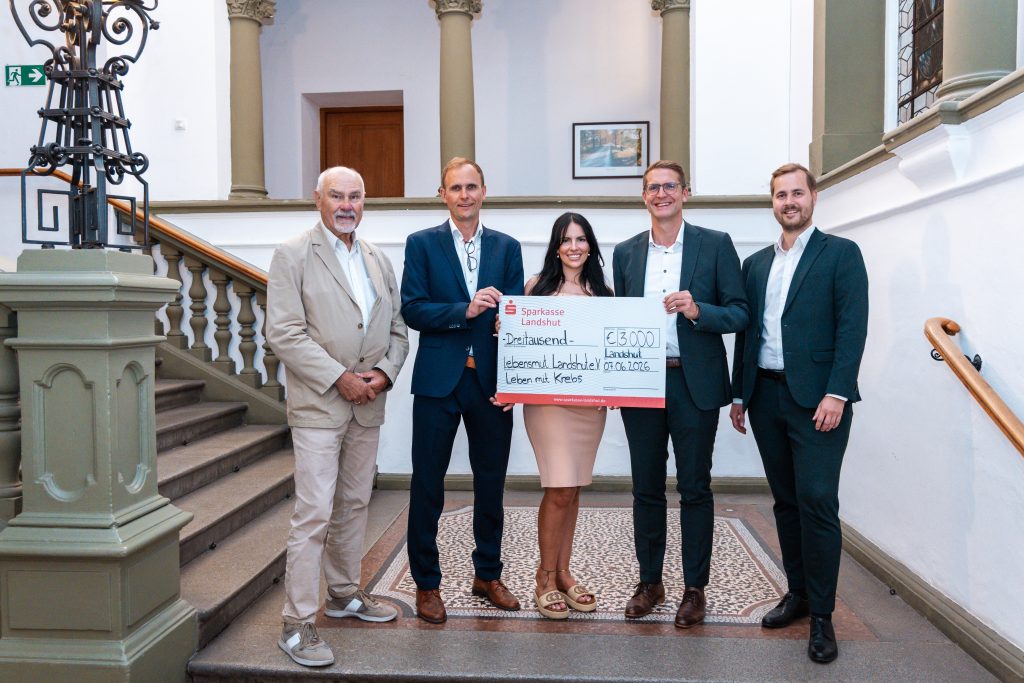 Five adults in formal attire stand on a grand staircase, presenting a large ceremonial check for €3,000 to a charitable cause.